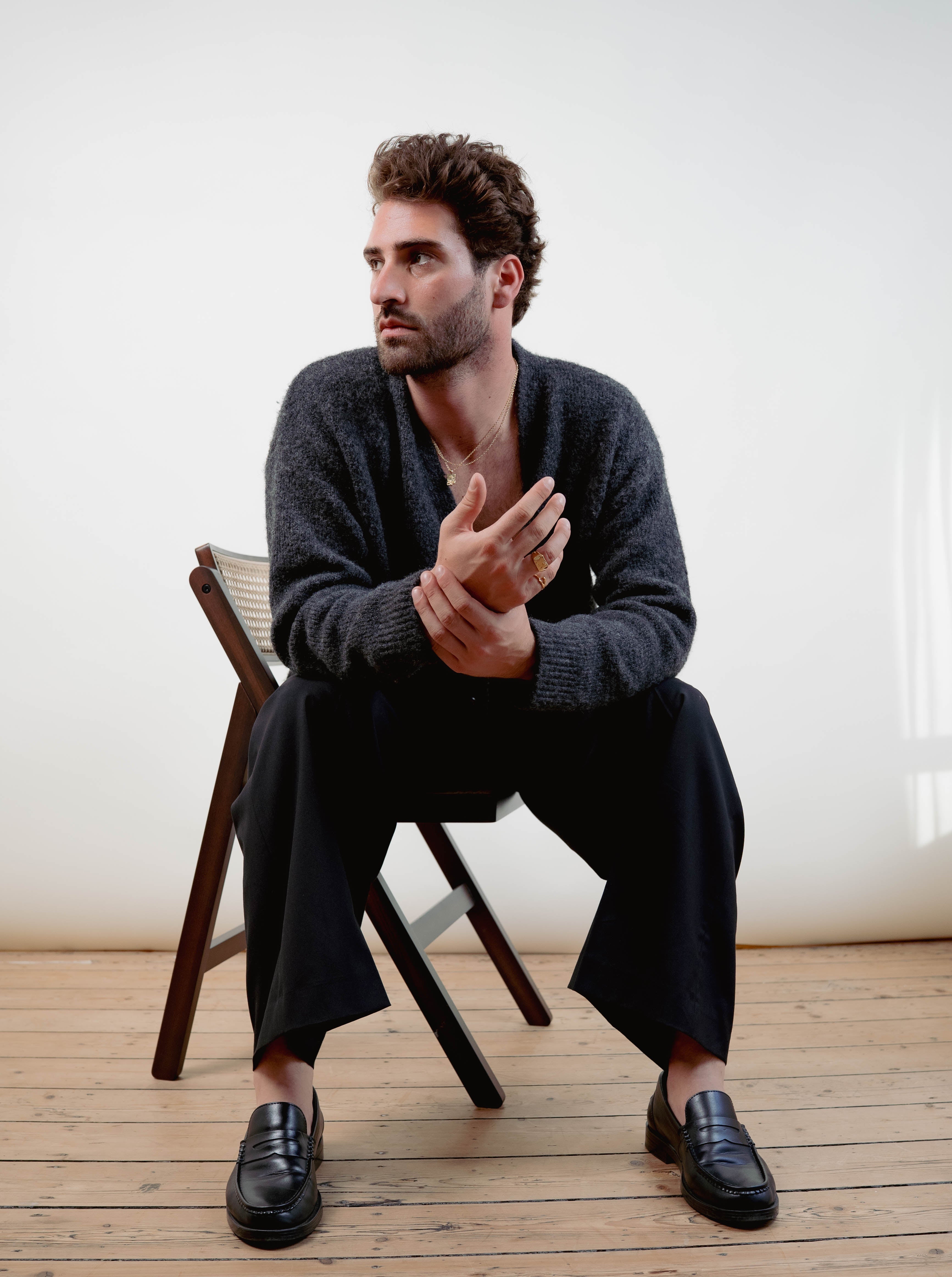 Man sitting on a wooden chair in a minimalistic room wearing EXCITÀRE jewelry