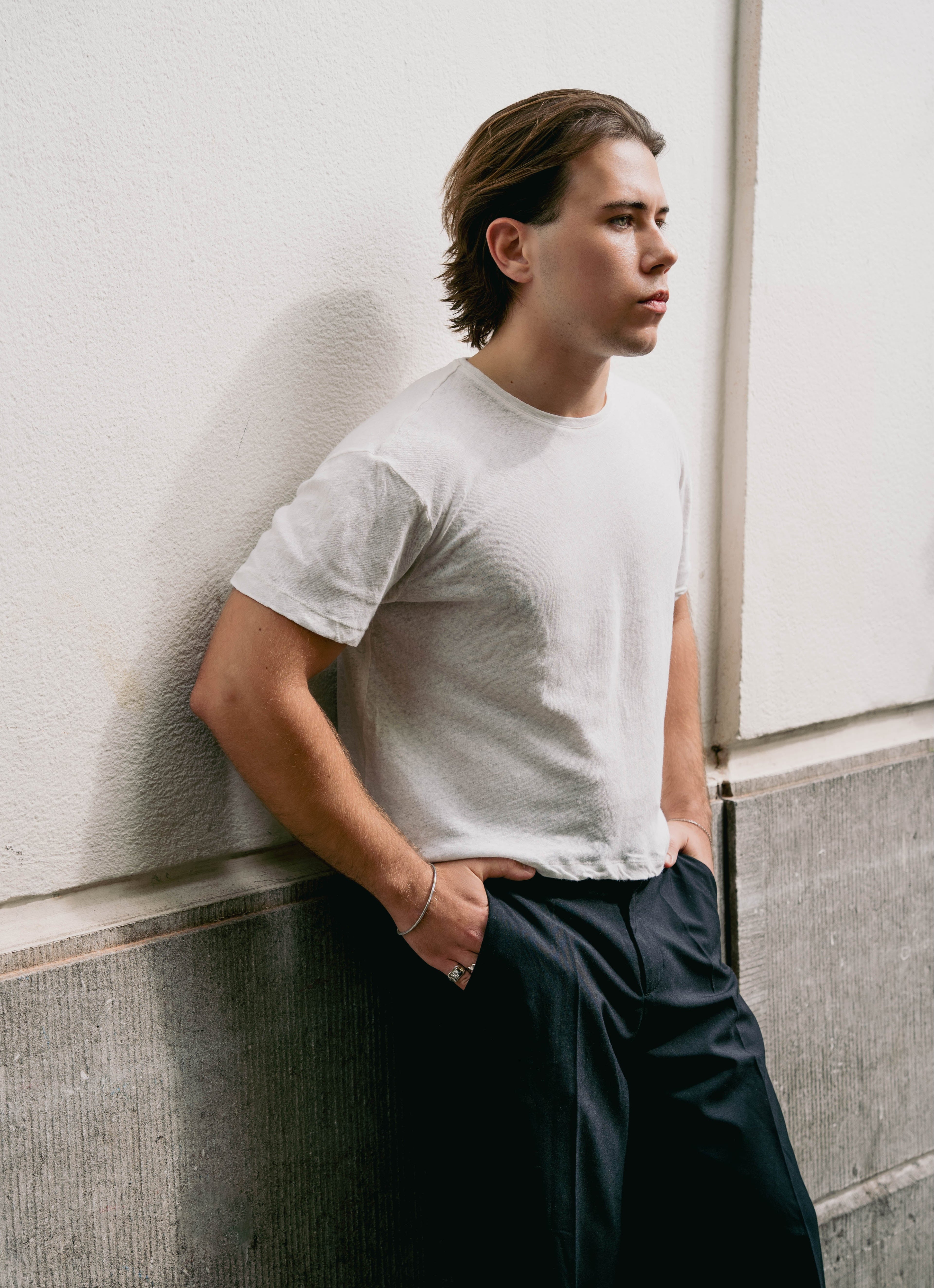 Man wearing a white t-shirt and dark pants leaning against a wall. Wearing EXCITÀRE jewelry