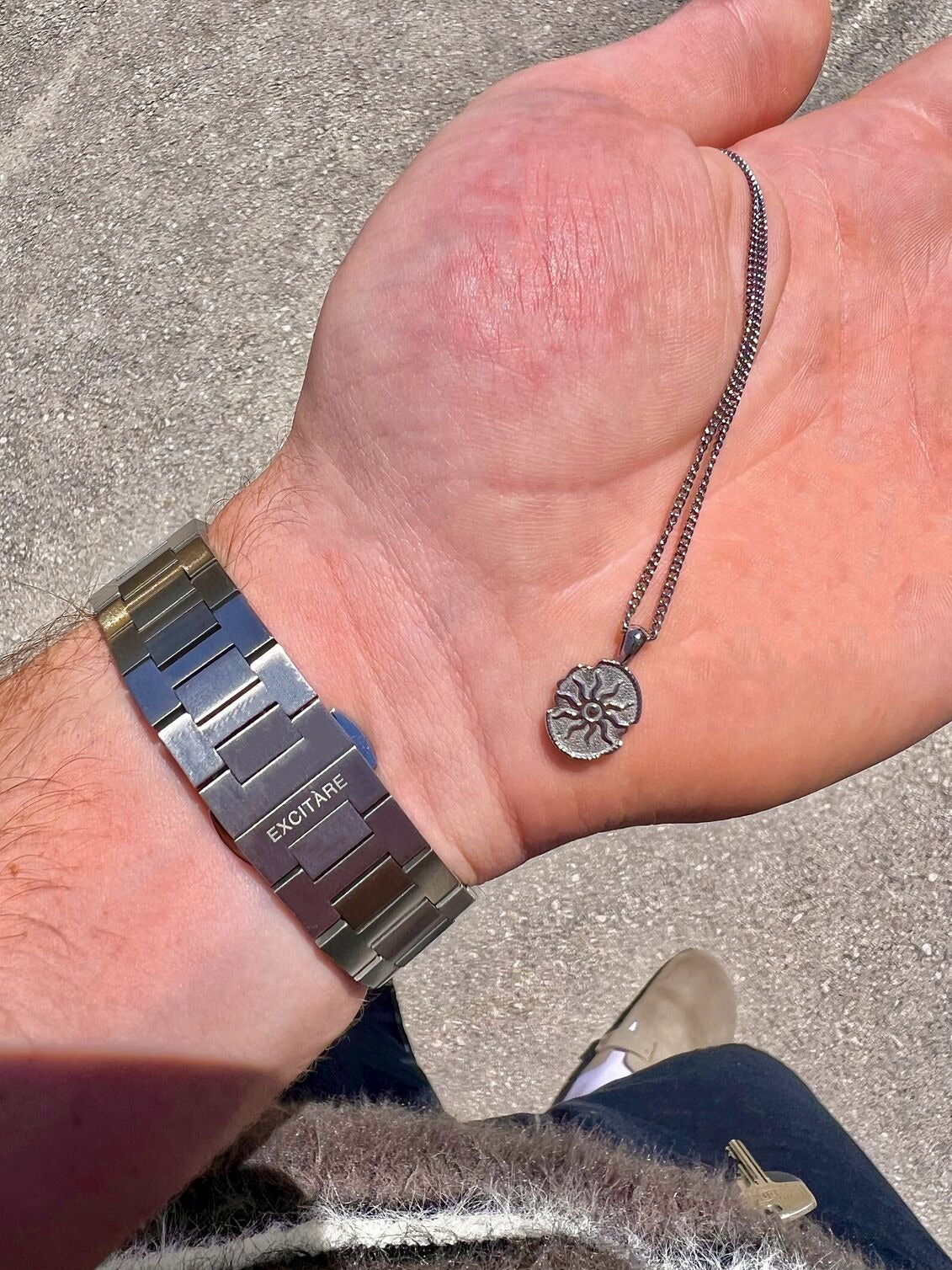 Close-up of a man's hand holding a silver EXCITÀRE Eclipse necklace, wearing a stainless steel EXCITÀRE watch on the wrist.