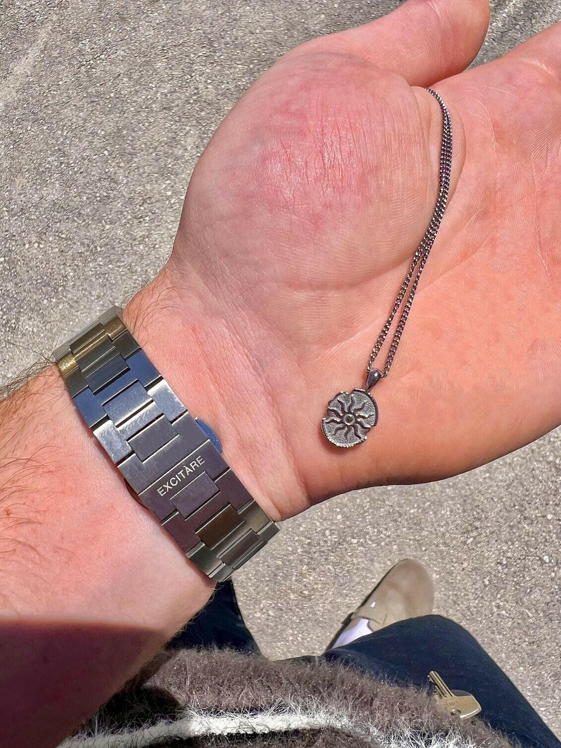 Close-up of a man's hand holding a silver EXCITÀRE Eclipse necklace, wearing a stainless steel EXCITÀRE watch on the wrist.