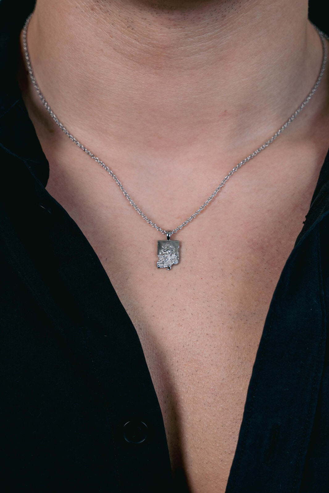 Close-up of a man wearing the EXCITÀRE Flower Necklace in silver, featuring a raw-edged rectangular pendant with a sculpted floral emblem, styled with an open black shirt.