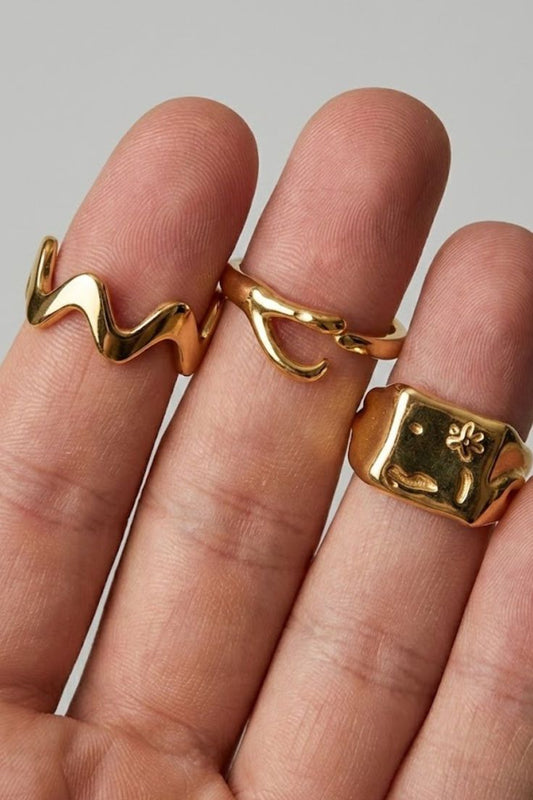 
  Close-up of the Ellis Ring Gold Extra ring stack, showing layered minimalist gold rings including a wave design, an open curve ring, and a bold signet ring, worn on the hand.
