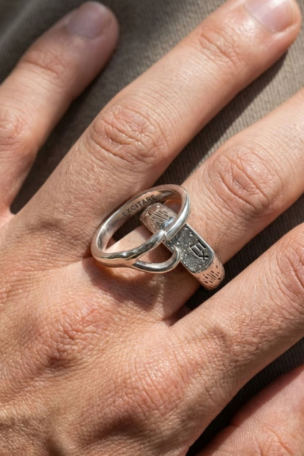 Close-up of the Excitare Ring Extra in silver, showing the engraved EXCITARE logo and textured details, worn on the hand in natural light.