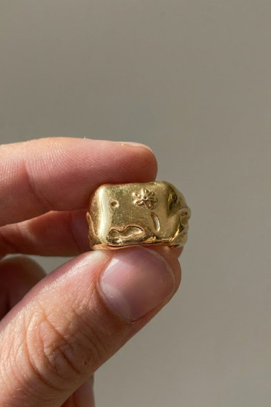 
  Close-up of the Flower Ring Gold Extra, a bold minimalist gold signet ring with engraved flower detail, held between fingers.
